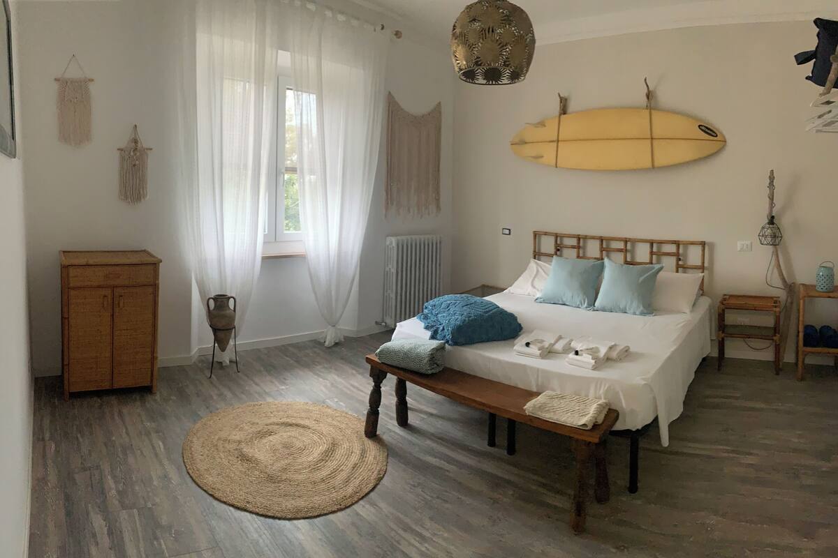 A bedroom with a bed, bedside tables, a wooden bench, a woven rug, a surfboard decoration, and a macramé wall hanging.