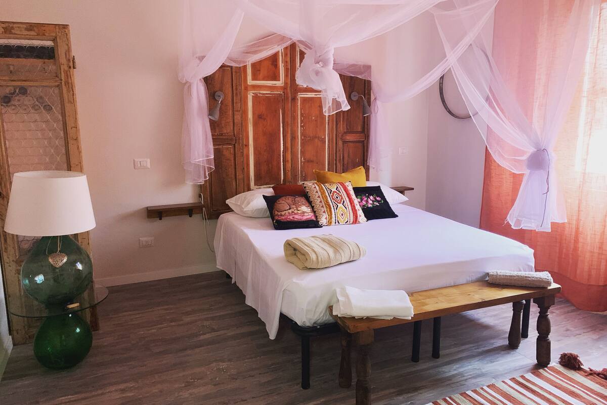 A bedroom with a bed, a bedside table, a lamp, a bench, and a wooden door.