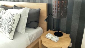 Una cama bien hecha con un cabecero, una mesita de noche con una lámpara y una pequeña mesa redonda con un objeto doblado encima.