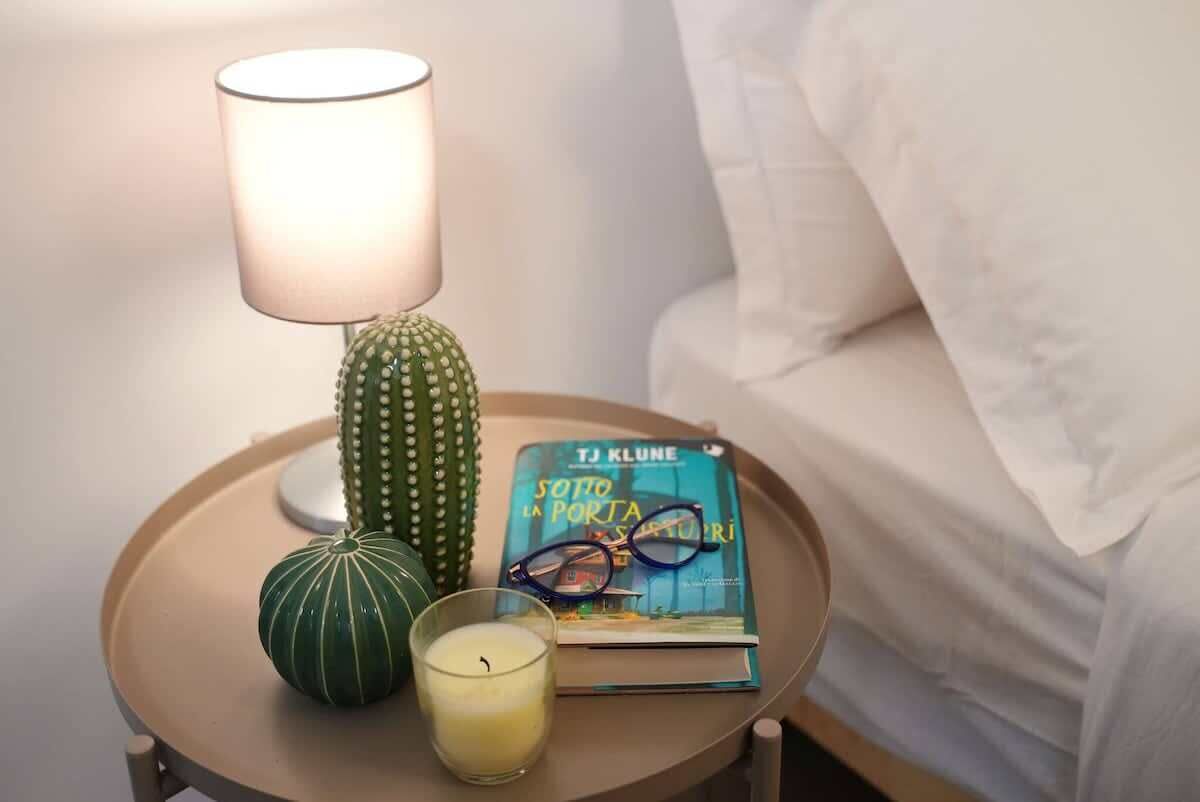Una mesita de noche con una lámpara, un libro, un par de gafas y un pequeño objeto verde.