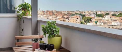 A balcony with a wooden bench, potted plants, and a view of a cityscape.