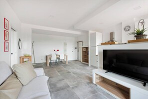 Un salon moderne doté d’un téléviseur à écran plat, d’un canapé, d’un coin repas avec une table et des chaises, et d’un coin cuisine avec des placards et une plante.