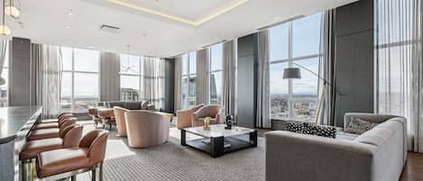 A modern hotel room with a bar, seating area, and large windows offering city views.