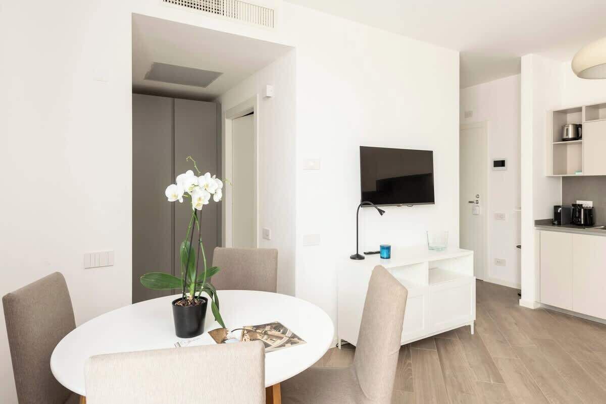 A modern living room with a round dining table, a flat-screen TV mounted on the wall, and a kitchen area with white cabinetry.