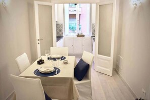 Un coin repas avec une table dressée pour quatre personnes, des chaises blanches et une vue sur un balcon, accessible par une porte ouverte.