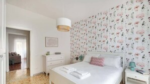 Une chambre à coucher comprenant un lit, une table de chevet, une lampe, une commode et un mur recouvert d’un papier peint à motifs floraux.