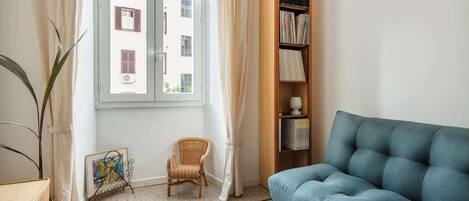 A room with a blue sofa, a wicker chair, a bookshelf, and a window with curtains.