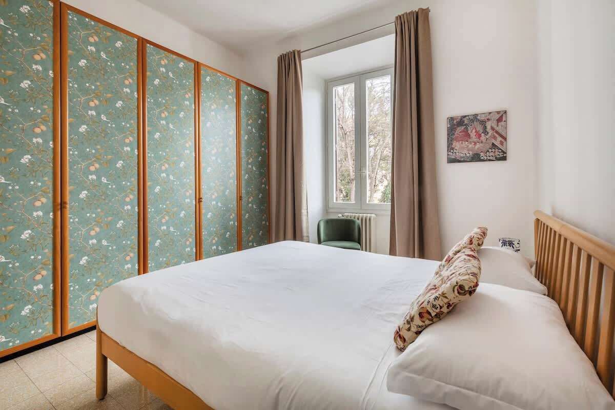 Ein Schlafzimmer mit einem Bett, einem grünen Sessel, einem Kleiderschrank und einem Fenster mit Vorhängen.