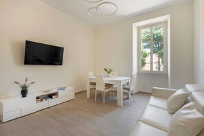 Uma sala de estar moderna com um sofá branco, uma mesa de jantar com cadeiras, uma TV fixada na parede e uma janela com vista para árvores.