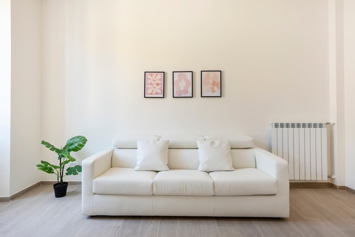 Ein minimalistisch eingerichtetes Wohnzimmer mit einem weißen Sofa, drei gerahmten Kunstwerken, einer Topfpflanze und einer Heizung.