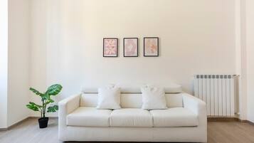 Ein minimalistisch eingerichtetes Wohnzimmer mit einem weißen Sofa, drei gerahmten Kunstwerken, einer Topfpflanze und einer Heizung.