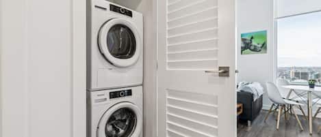 A laundry room with two stacked front-loading washers and dryers, a white door with horizontal slats, and a view of a cityscape through a large window.