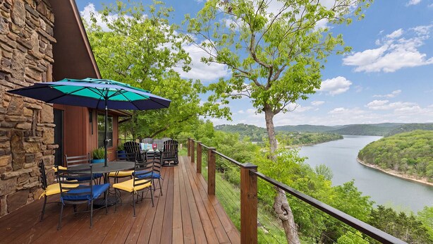 Eine Holzterrasse mit Gartenmöbeln und einem Tisch, mit Blick auf einen Fluss und ein bewaldetes Gebiet.