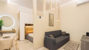 Une pièce moderne et bien éclairée, avec un canapé gris, une petite table et un lit doté d’une tête de lit en bois.