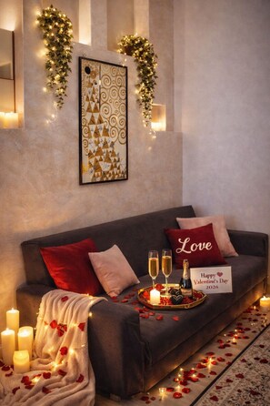 Ein gemütliches Wohnzimmer mit einem dunkelgrauen Sofa, roten und weißen Kissen, einem gerahmten Kunstwerk und einer Liebesherz-Dekoration, darunter Kerzen, Champagnergläser und ein Schild.