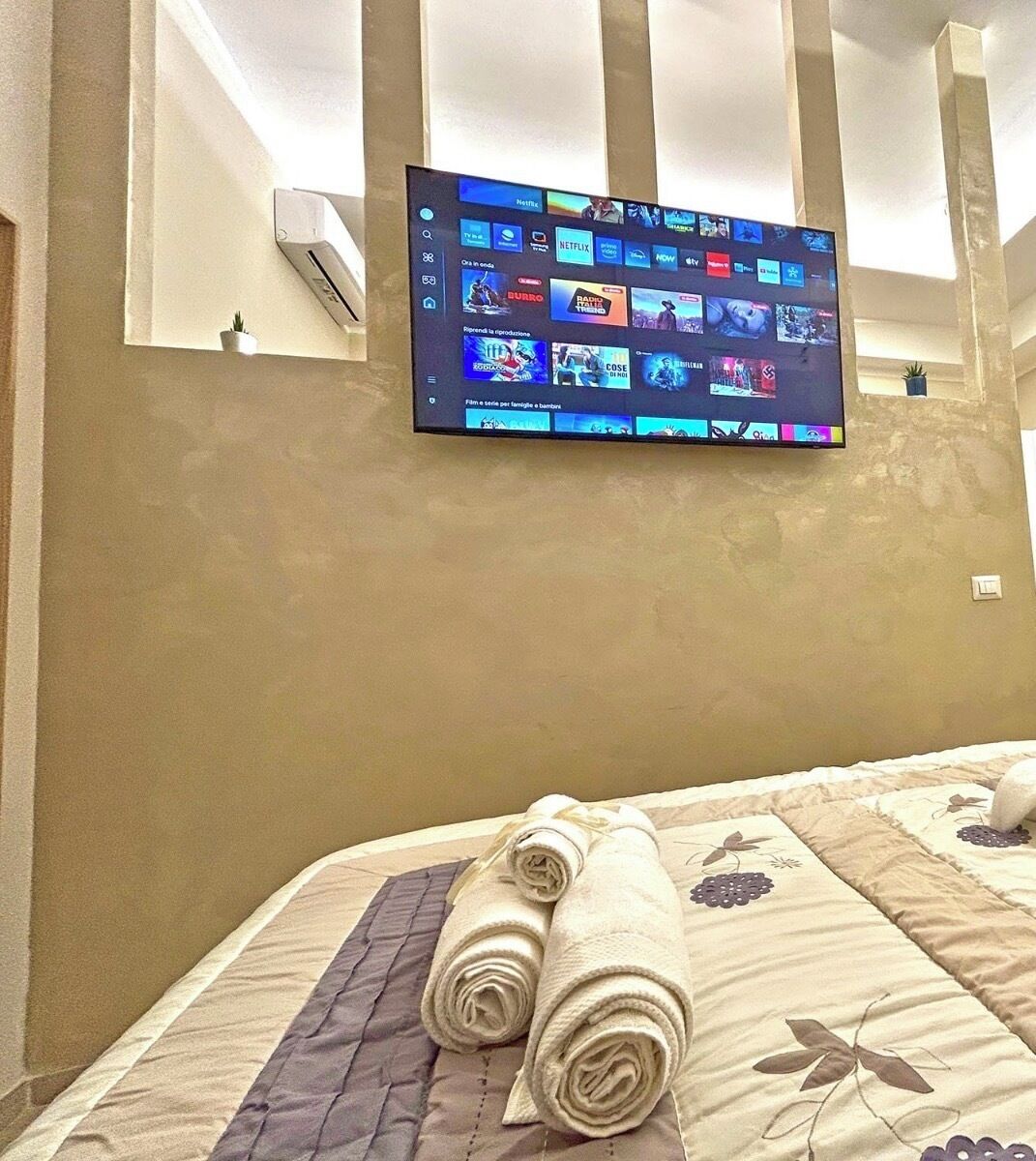 A hotel room with a bed, a TV mounted on the wall displaying a streaming service interface, and a small potted plant on the nightstand.