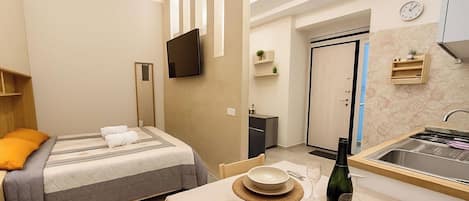 Habitación de hotel compacta con cama, mesita de noche y una pequeña cocina.