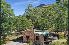 A stone building with a covered patio, surrounded by dense trees and a mountain in the background. - Halls Gap House,  Grampians (Halls Gap)