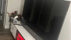 Una stanza con una grande TV a schermo piatto montata a parete, un mobile bianco con cassetti e un tavolino con oggetti decorativi.
