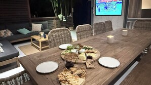 A dining area with a long wooden table set for a meal, surrounded by wicker chairs, with a TV displaying a rugby match in the background. - Cowan Lodge Cowan Cowan (Cowan Cowan)