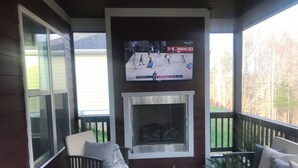 A covered porch with a fireplace, seating area, and a television displaying a basketball game.