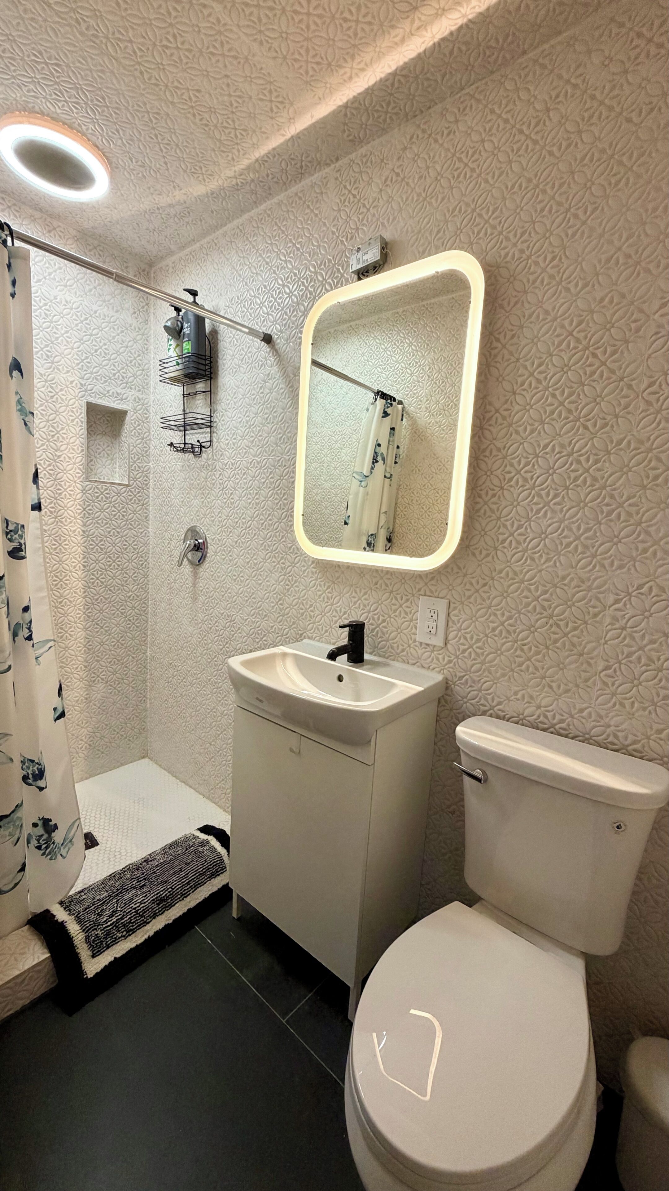 A bathroom with a shower, sink, toilet, and a mirror with a light.
