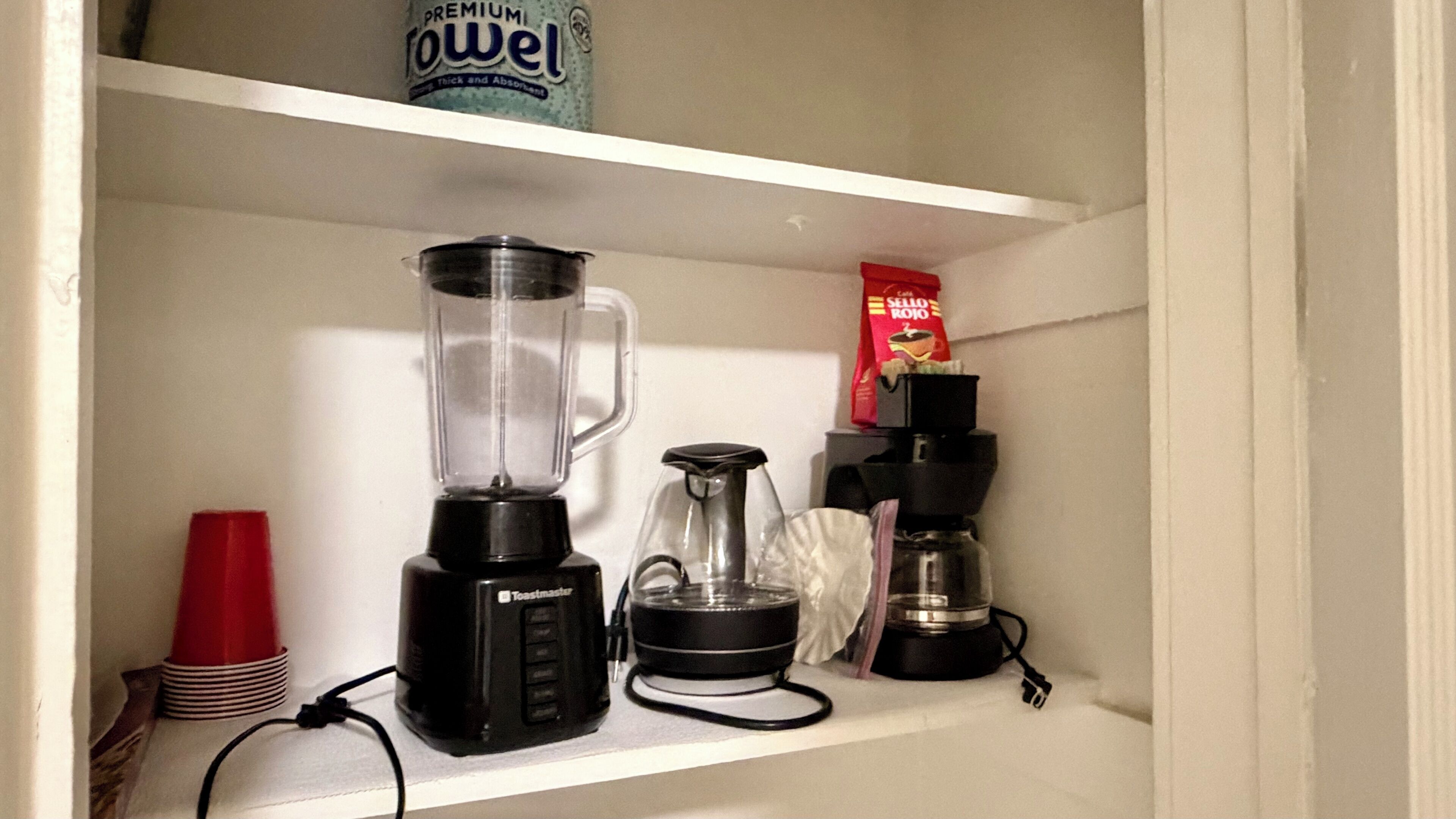 A kitchen shelf with a blender, a coffee maker, a red cup, and a box of Sello Rojo.