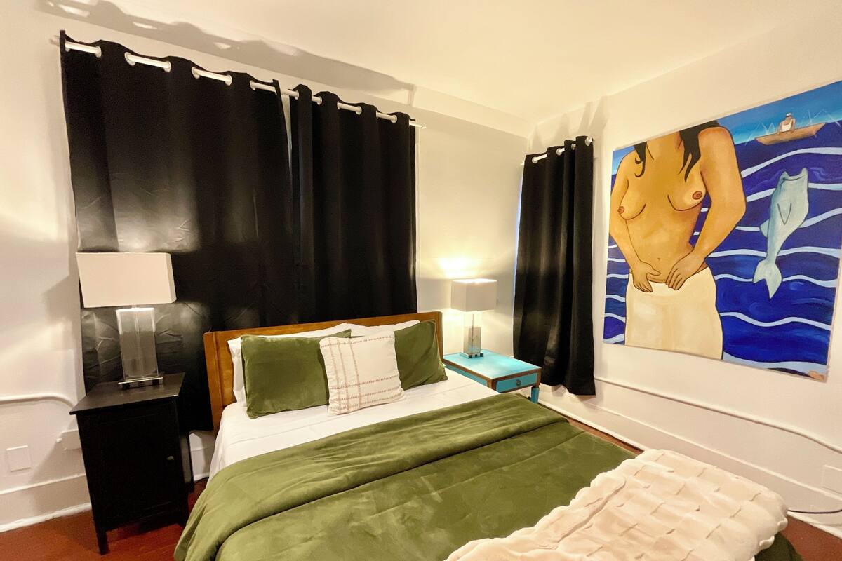 A bedroom with a bed, two bedside tables, a nightstand, a painting, and black curtains.