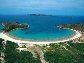 An aerial view of a coastal area with a curved bay, sandy beach, and surrounding greenery.