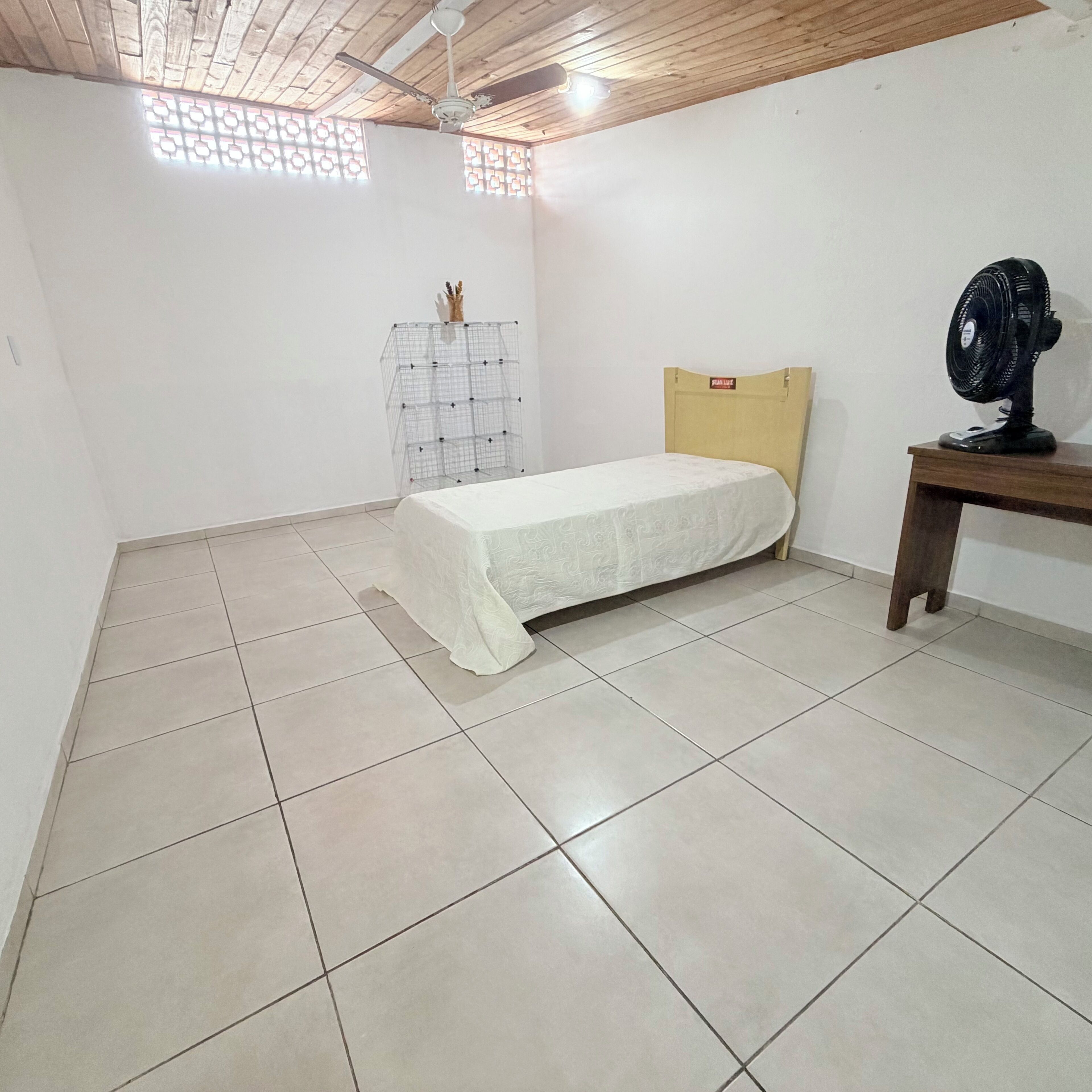 Une chambre à coucher comprenant un lit, un ventilateur de plafond, une petite table et une fenêtre à carreaux.
