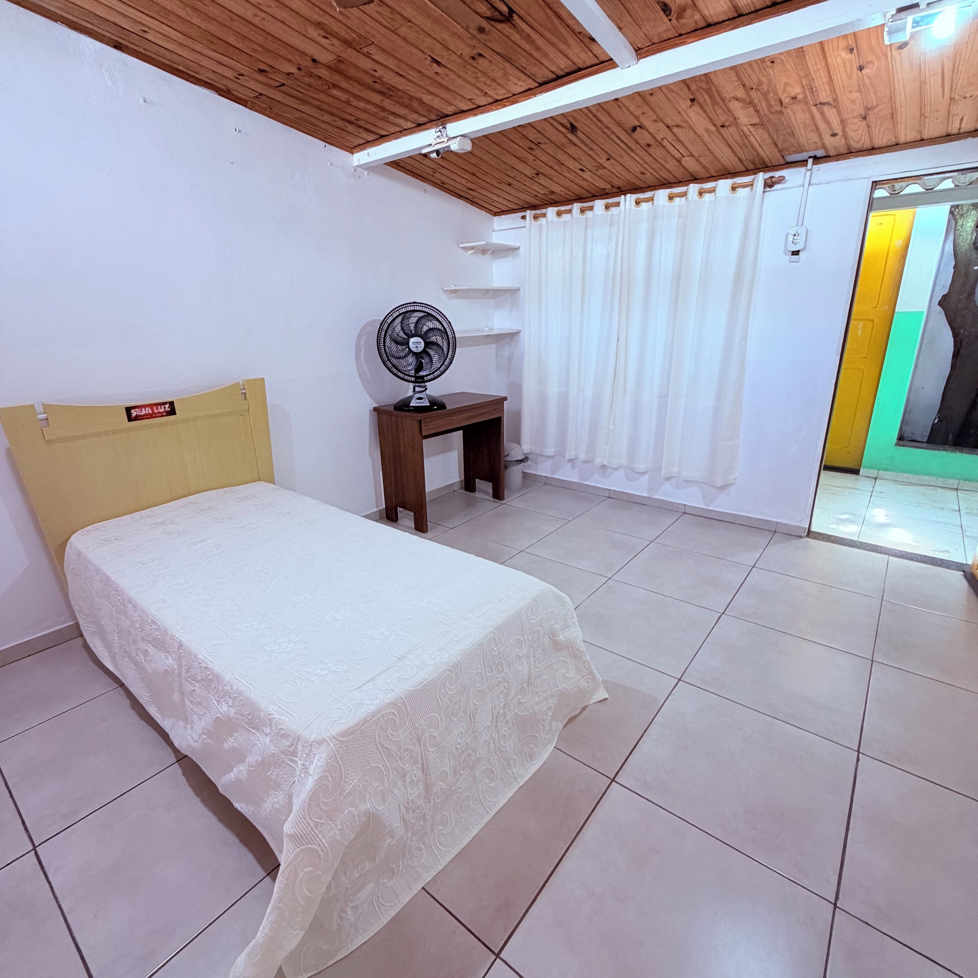 Quarto com cama, ventilador, teto de madeira e porta que dá para outro cômodo.