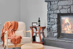 A cozy living room with a stone fireplace, a comfortable armchair with a throw blanket, and a small wooden table with a bottle and a framed picture. - Spacious Poconos Retreat for Families & Groups (Tobyhanna)