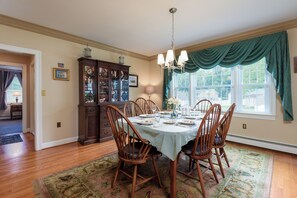 Comedor con una mesa dispuesta para cuatro personas, rodeada de sillas de madera, un candelabro y un amplio ventanal con cortinas verdes.
