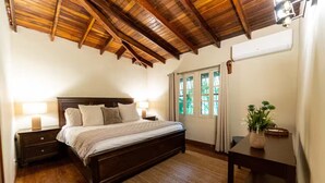 Camera da letto con soffitto in legno, un letto con molti cuscini, un comodino con una lampada, una finestra con tende e un tavolino con una pianta.