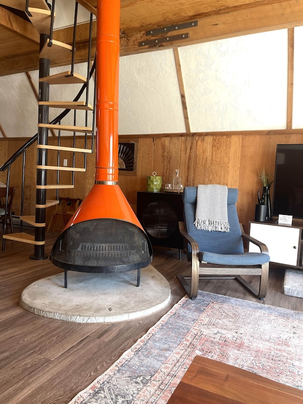 A modern living room with a wooden staircase, a round orange heater, a blue armchair, and a flat-screen TV.