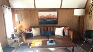 A cozy living room with a brown leather sofa, a wooden coffee table, and a painting above it. There are two woven poufs on the floor, a lamp on a side table, and a chair in the corner.