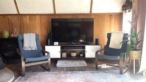 A living room with a wooden ceiling, two blue armchairs, a TV on a white cabinet, and a patterned rug.