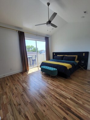 A bedroom with a bed, a ceiling fan, wooden flooring, and a view of the outdoors.