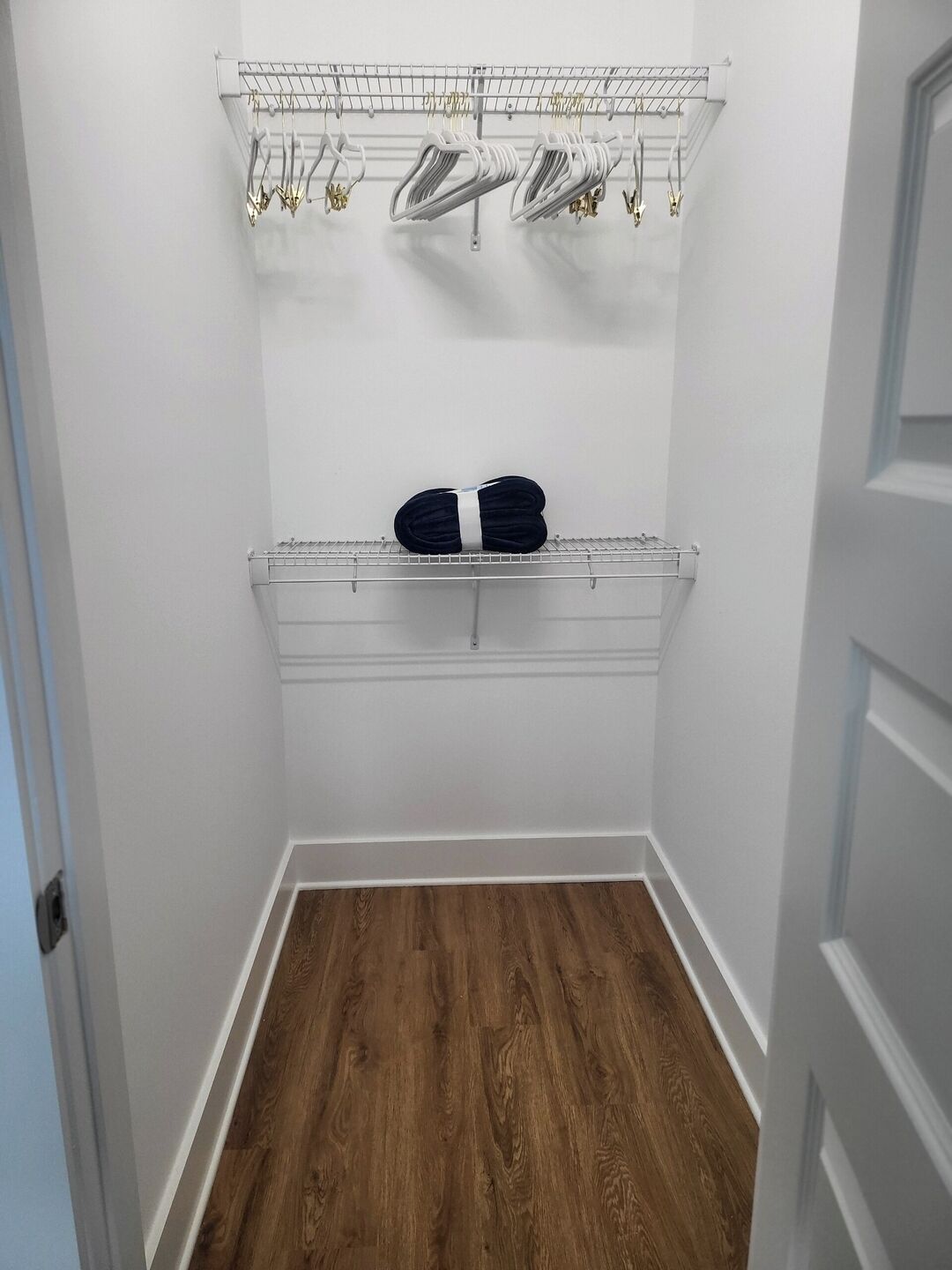 A small, narrow hallway with a wooden floor and white walls. There is a metal rack with hangers and a folded black item on a shelf.