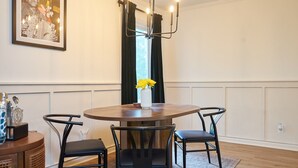 A dining area with a round wooden table, two black chairs, a vase with yellow flowers, a framed picture on the wall, and a modern chandelier. - Cozy 3-Bedroom House in Quaint Douglasville (Douglasville)