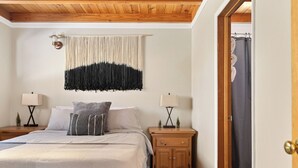 A bedroom with a bed, two bedside tables, a wall hanging, and a door to a bathroom.