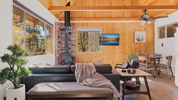 A cozy living room with a fireplace, a dining area, and large windows offering views of trees and a lake.