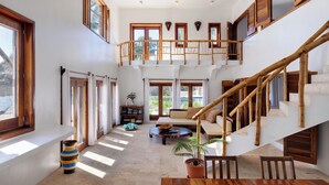 Un'ampia zona giorno con soffitto alto, ampie finestre, una scala in legno e un angolo salotto con divano e tavolino.