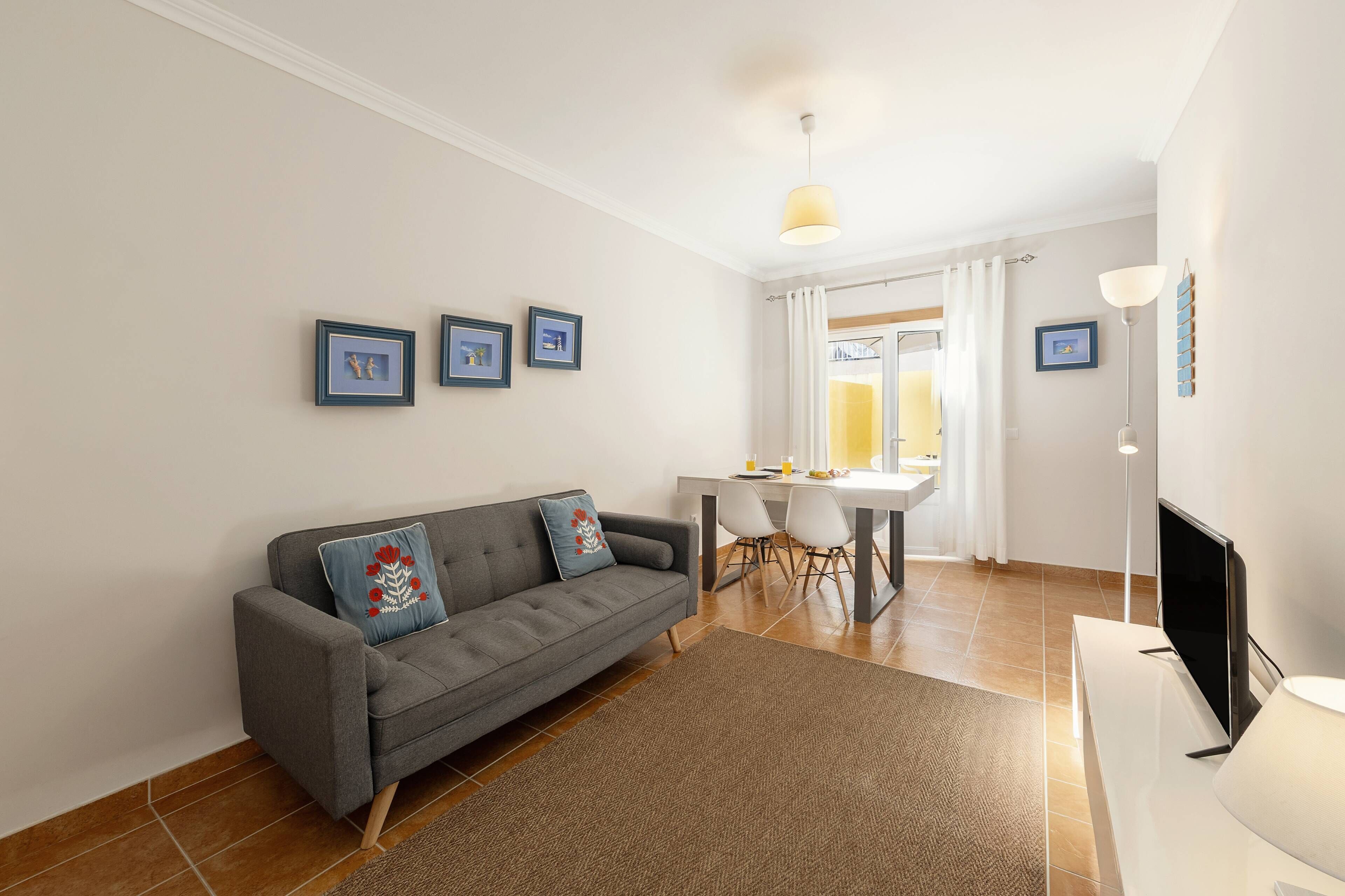 Una sala de estar moderna con un sofá gris, una alfombra marrón, un televisor de pantalla plana y un comedor con mesa y sillas.