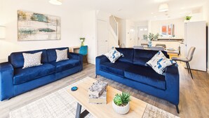 Un salon moderne avec deux canapés bleus, une table basse avec des livres et une plante, et un escalier menant à un espace repas.