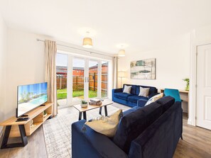 A modern living room with a large TV, a sofa, a coffee table, and a view of a garden through glass doors. - Rivoli Nightingale – 3 Bedroom Home, Free Parking, Central Derby & Station (Derby)