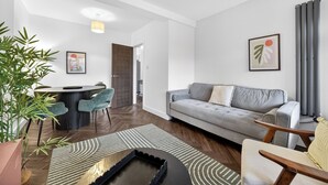A modern living room with a grey sofa, a dining area with a round table and chairs, and a large potted plant. - Amber Vista Apartments Cardiff (Cardiff)