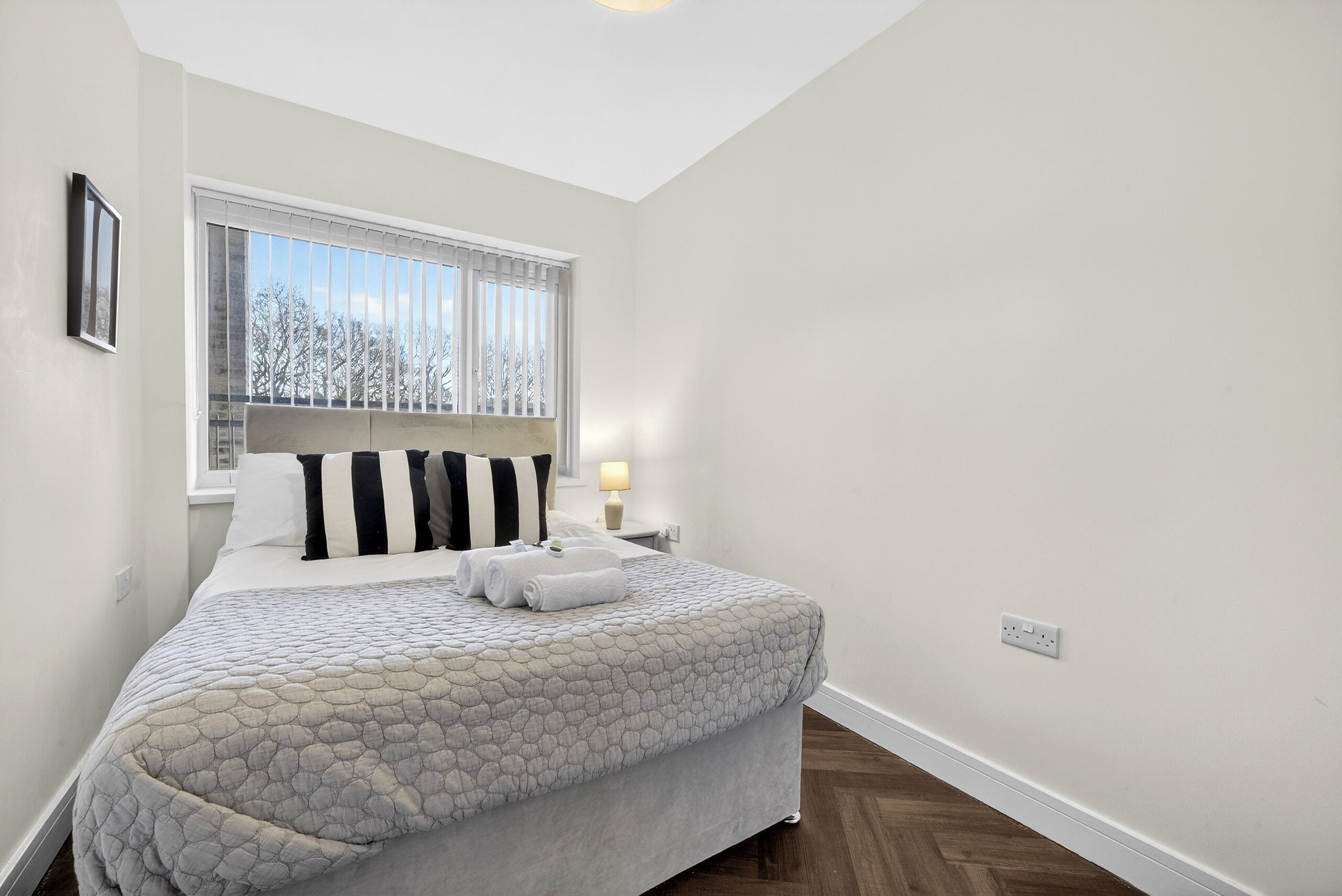 A neatly made bed with a patterned bedspread, a window with blinds, and a bedside lamp.