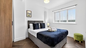 A bedroom with a bed, a green ottoman, a window with blinds, and a ceiling light. - Amber Vista Apartments Cardiff (Cardiff)