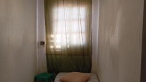 Une petite pièce comprenant un lit, un petit tabouret et une fenêtre ornée de rideaux légers.